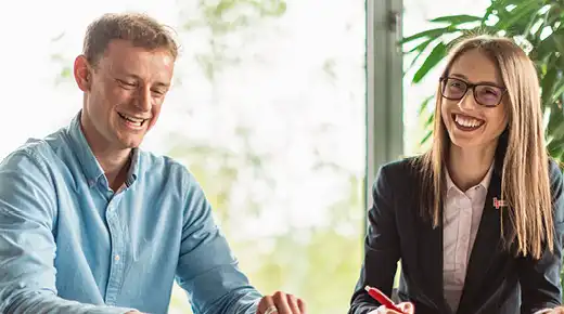 Two employees sit next to each other, talking & smiling. In the background, office plants can be seen.