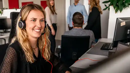 Woman with headset smilingly sits in an office, while participating in a video call.
