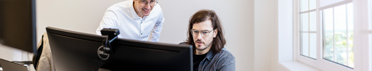 One colleague helping another, who is seated at a desk behind a computer