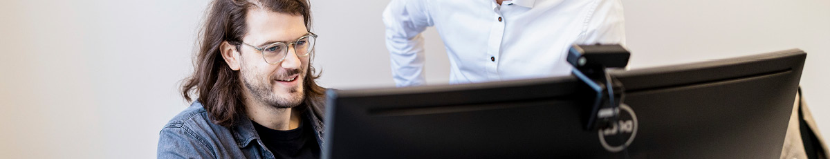 Two colleagues in an office discuss something on a computer screen in front of them.
