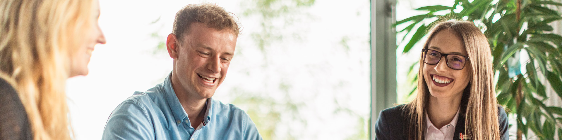 Three employees of Johner Institute are having a meeting in an office setting. All are laughing. In the background, a large window pane lets in natural light; plants can be seen.