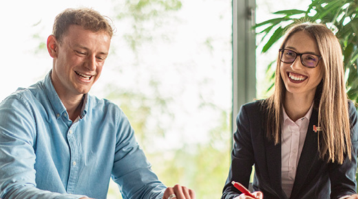Two colleagues sitting next to each other in an office talk and smile.