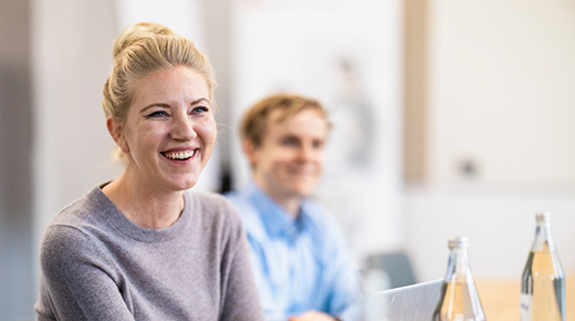 Woman in a conference room smiling at someone out of frame; her colleague in the background is also smiling.